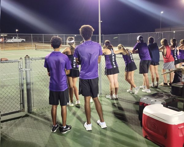 TIED UP. Members of the tennis team wait on Friday for senior Brayden Leinweber to finish his singles game. In an unusual twist, both players were struggling with cramping in the incredibly long match, but Leinweber was able to continue playing, winning the game and match. 